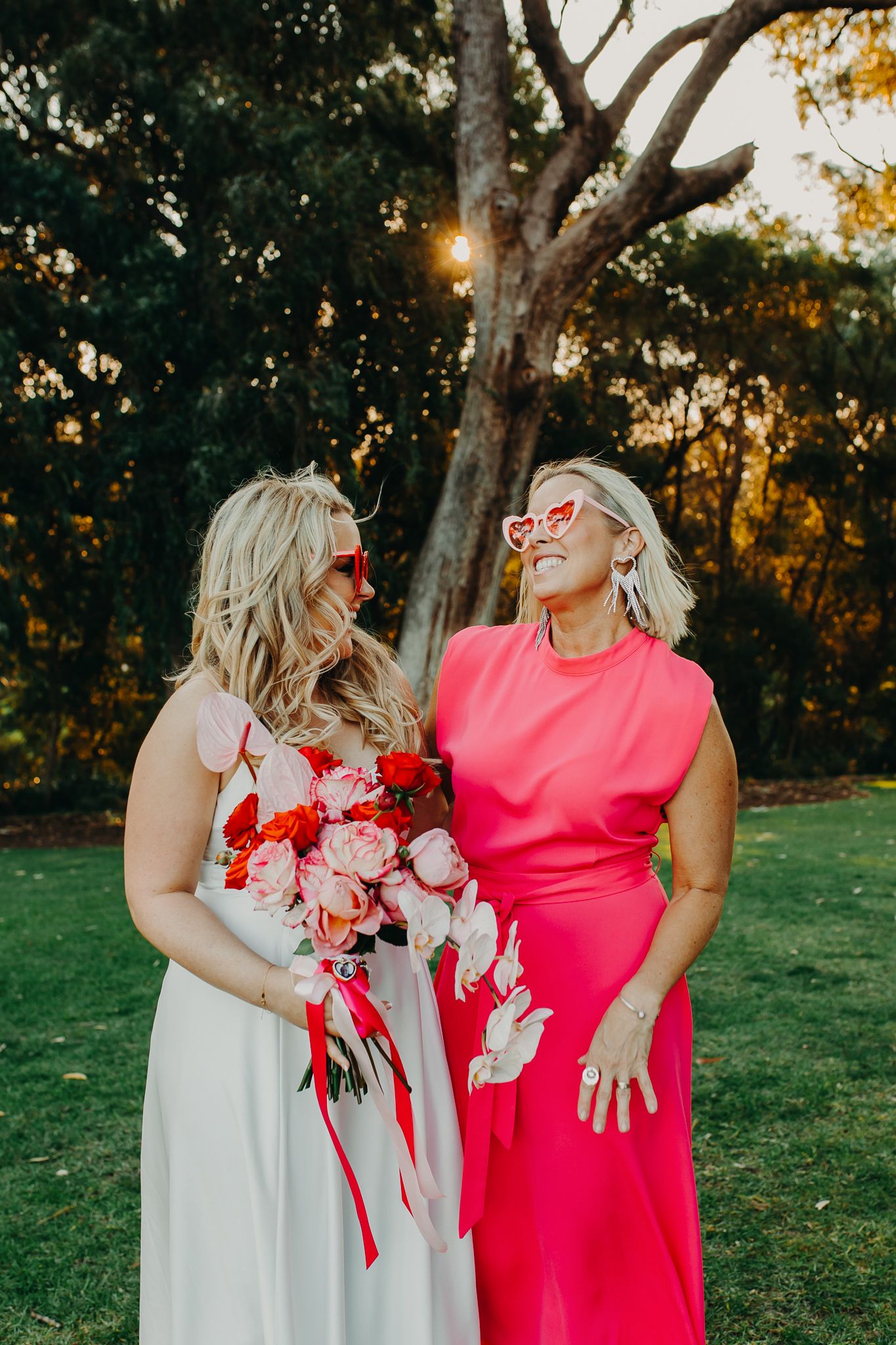 Two women in white and pink dresses laugh together while holding flowers at sunset.