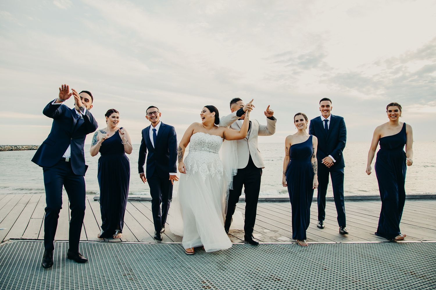 Wedding party poses together on dock with water and cloudy sky backdrop.