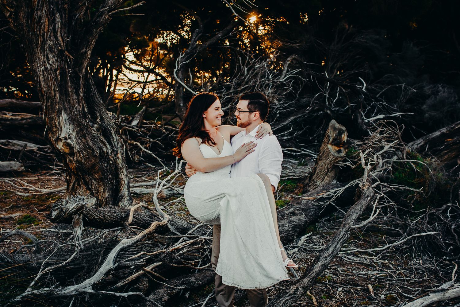 Couple embraces in romantic pose among twisted tree branches at sunset.