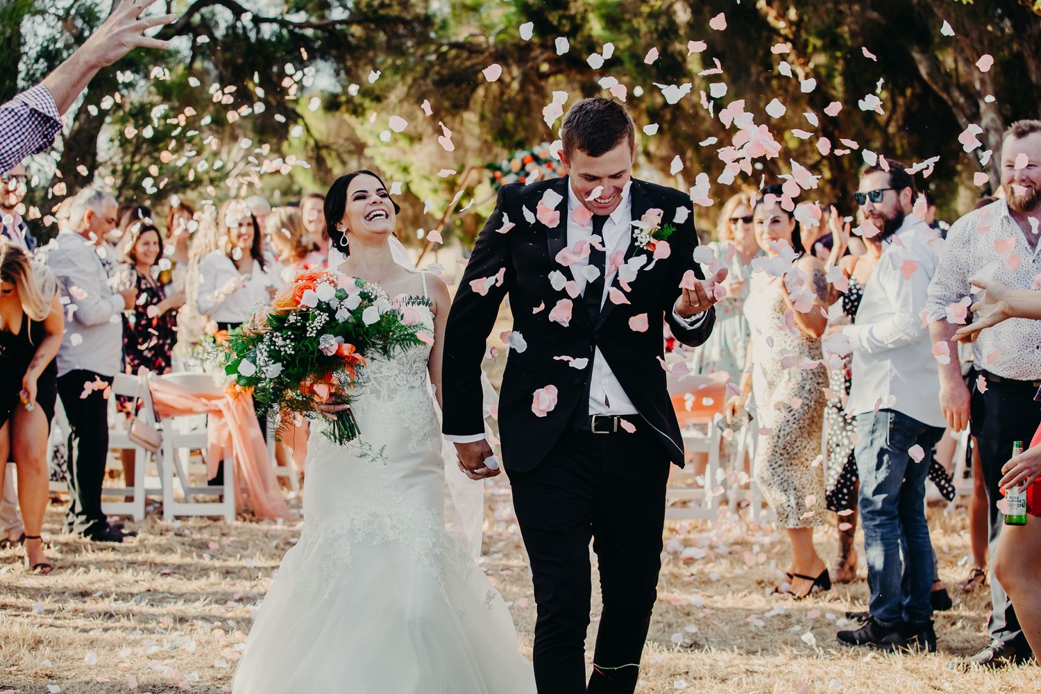 Newlyweds walk through shower of confetti on tree-lined path.