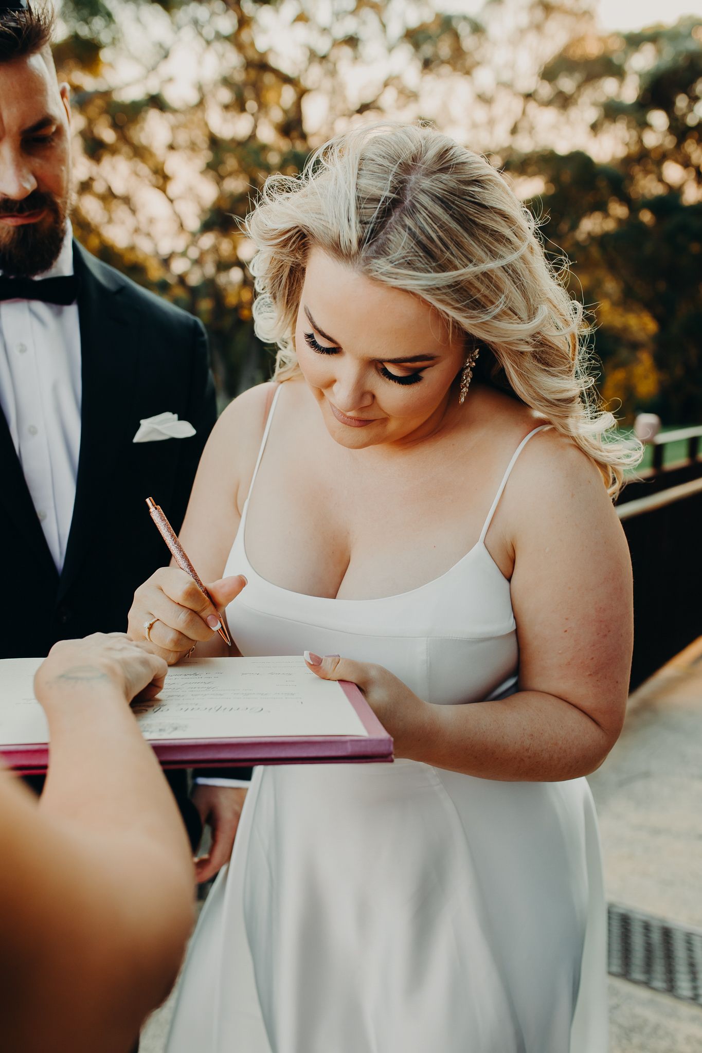 Woman in white dress signs document during outdoor ceremony at sunset.