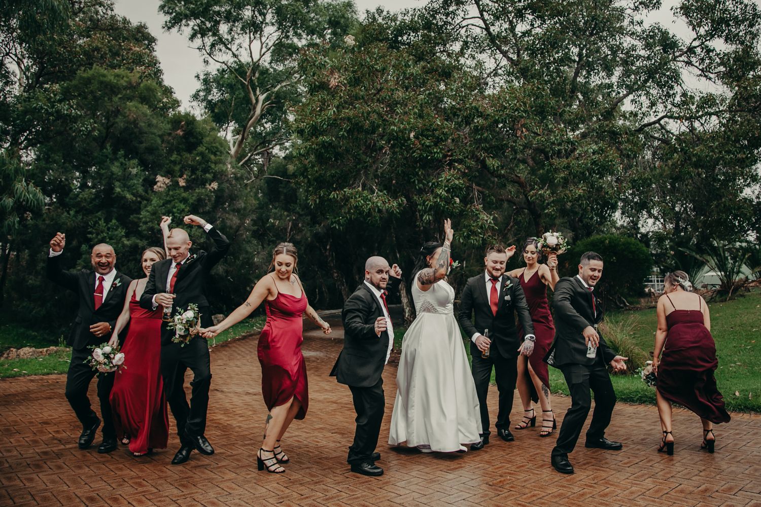 Wedding party dances together on a dirt path surrounded by trees during an outdoor celebration.