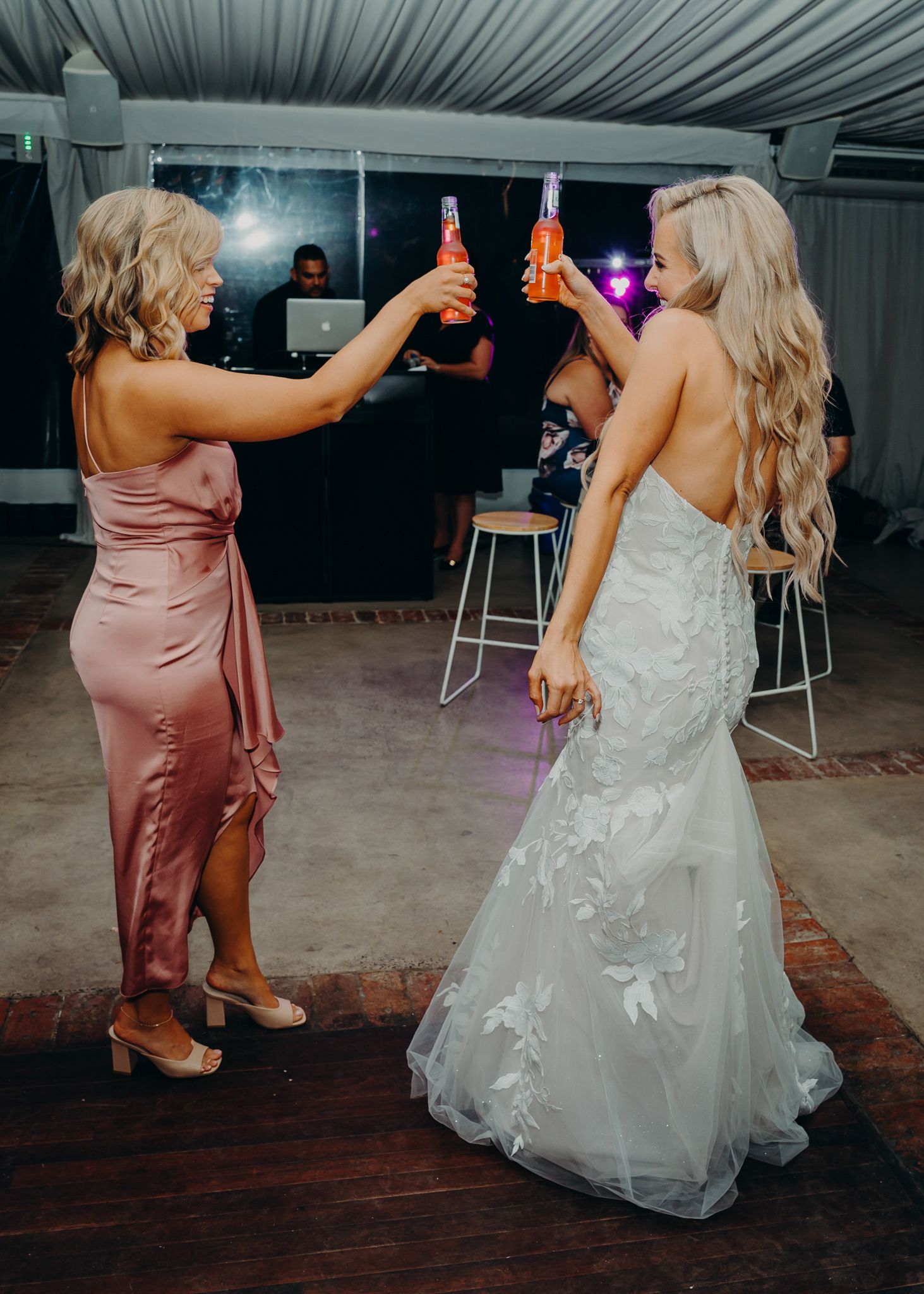 Two women in formal attire toast with bottles under a white tent at a celebration.