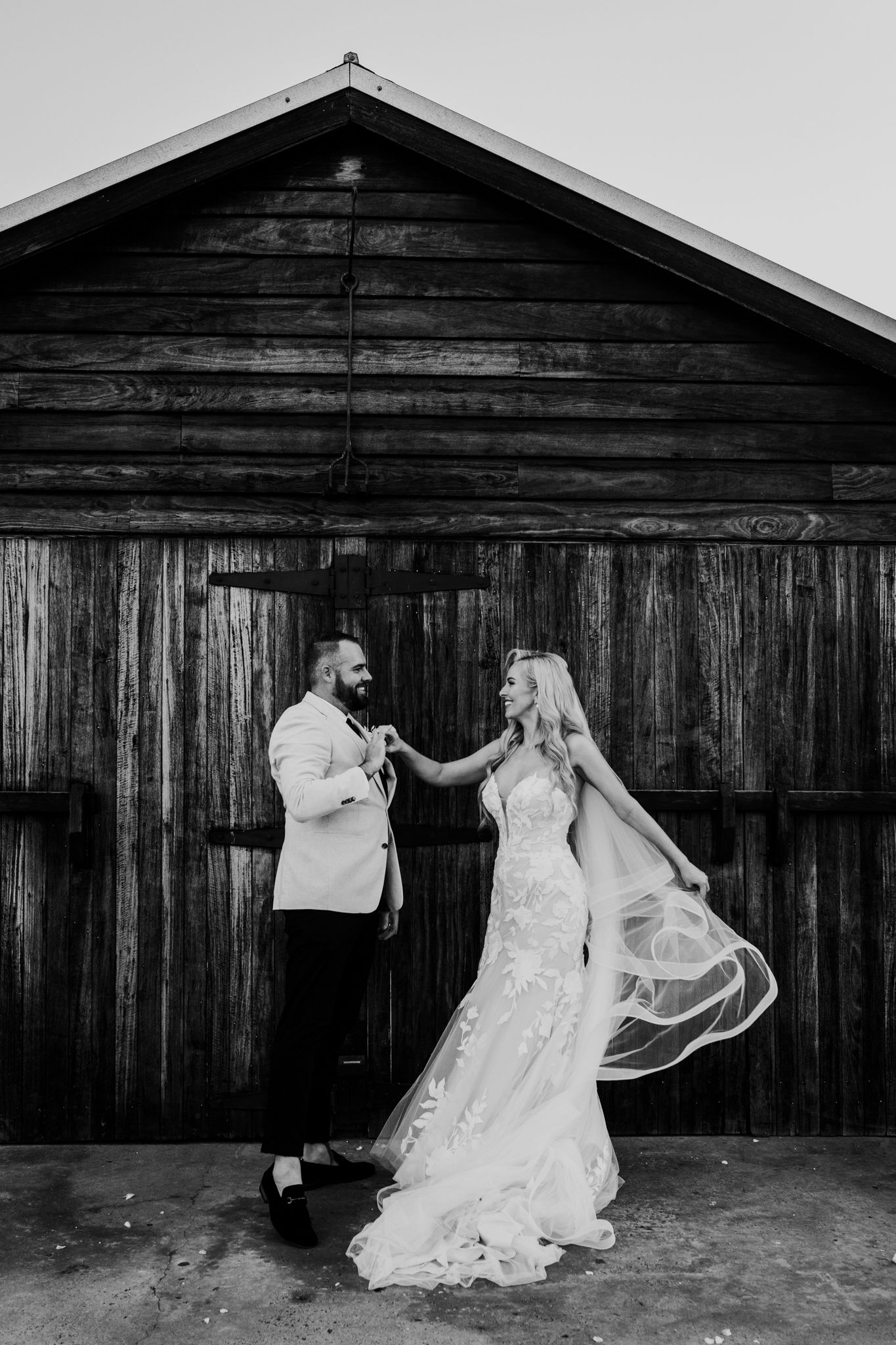 Black and white portrait of couple in wedding attire dancing by rustic wooden barn.