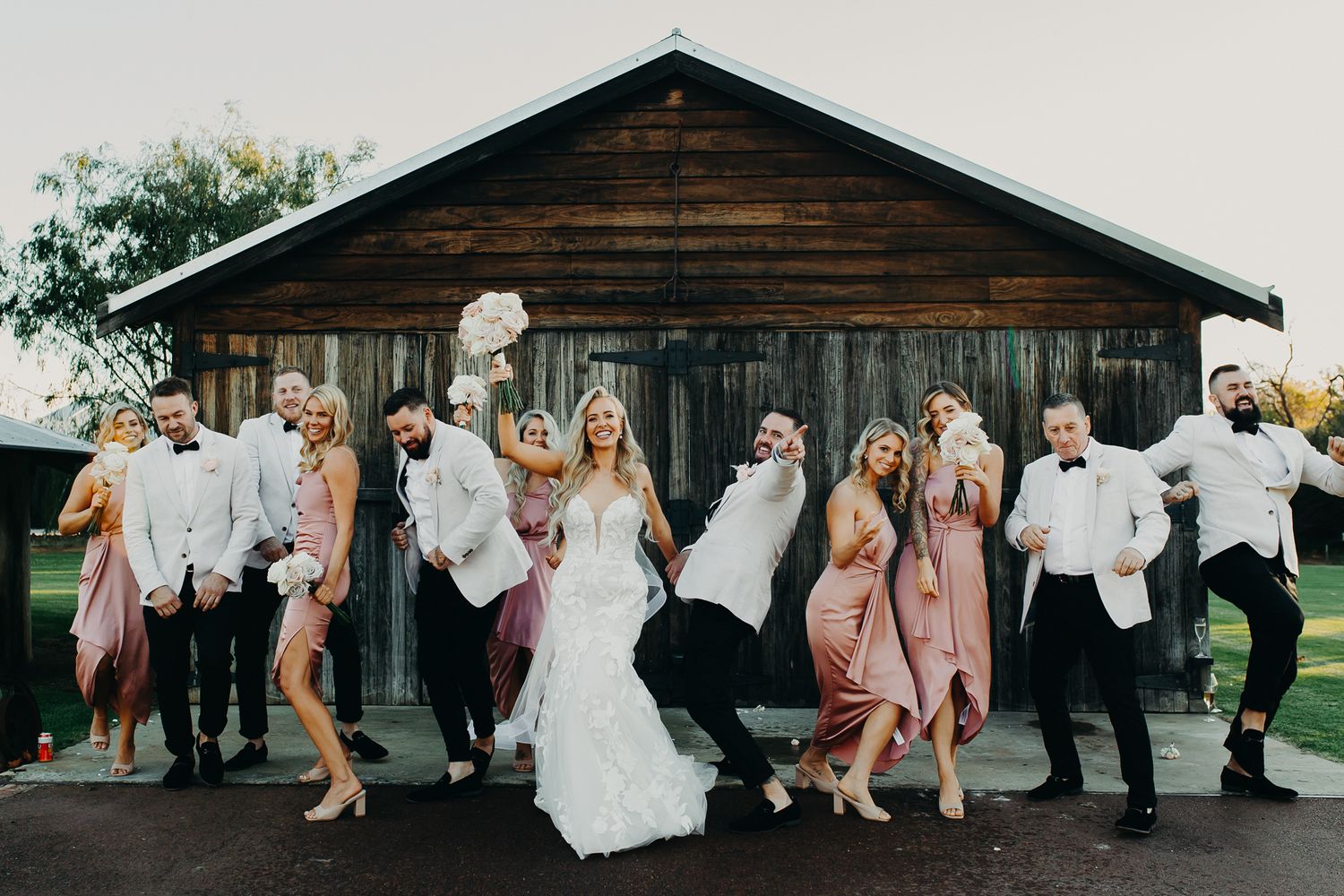 Wedding party poses energetically in front of rustic wooden barn.