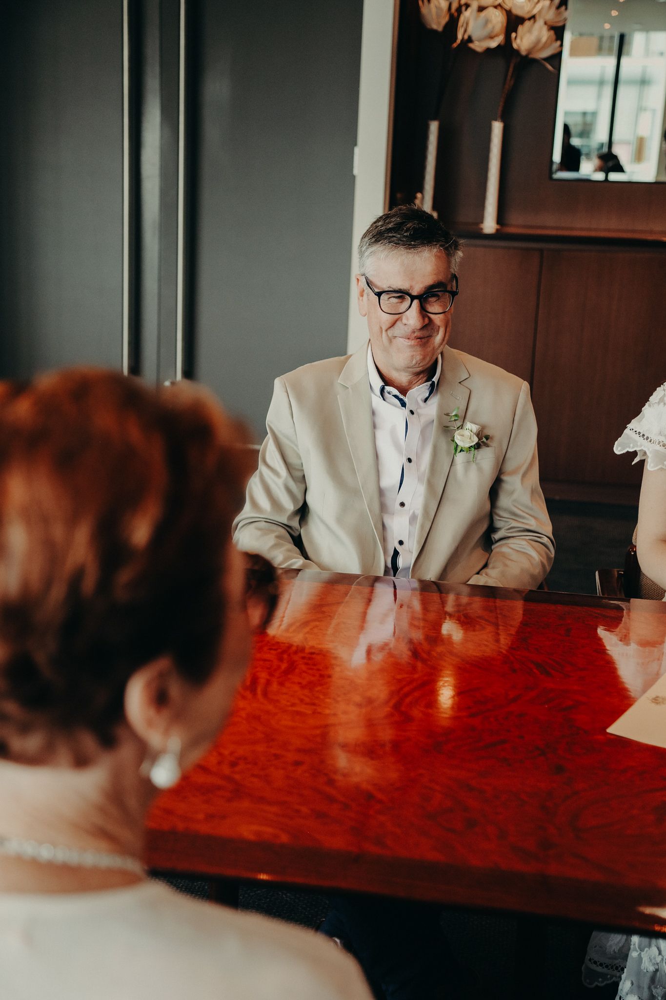 Person in beige suit with boutonniere sits at glossy red table during wedding event.