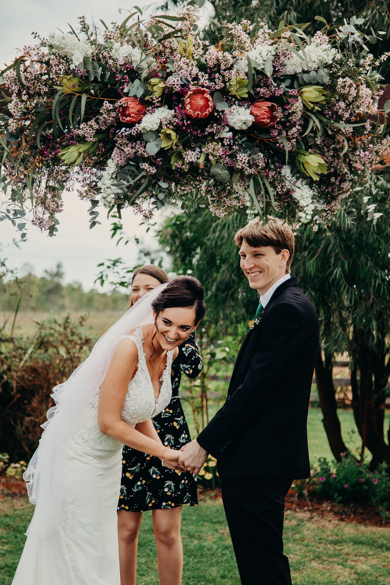 Wedding couple and guest share a laugh under stunning floral arch with pink and red blooms in outdoor garden setting.