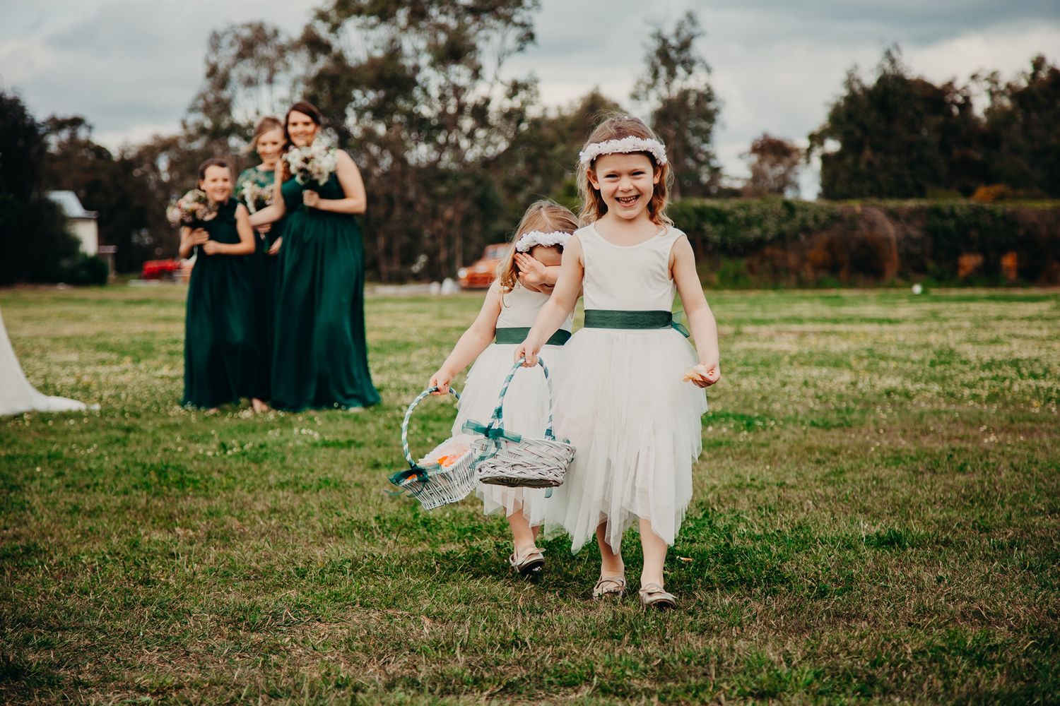 Flowergirls in white and emerald dresses walk across a grassy field during wedding ceremony.