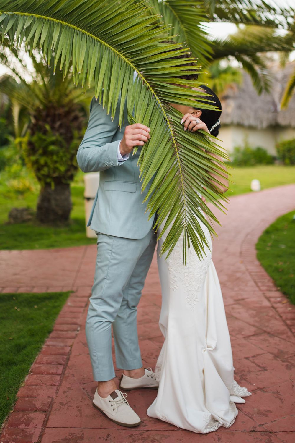 Couple playfully hiding behind a large palm frond on a brick pathway in a tropical garden setting.