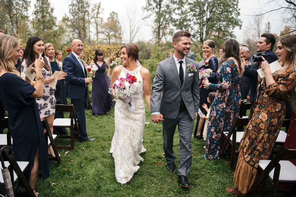 Wedding couple walks down outdoor aisle surrounded by celebrating guests on grassy lawn with trees in background.