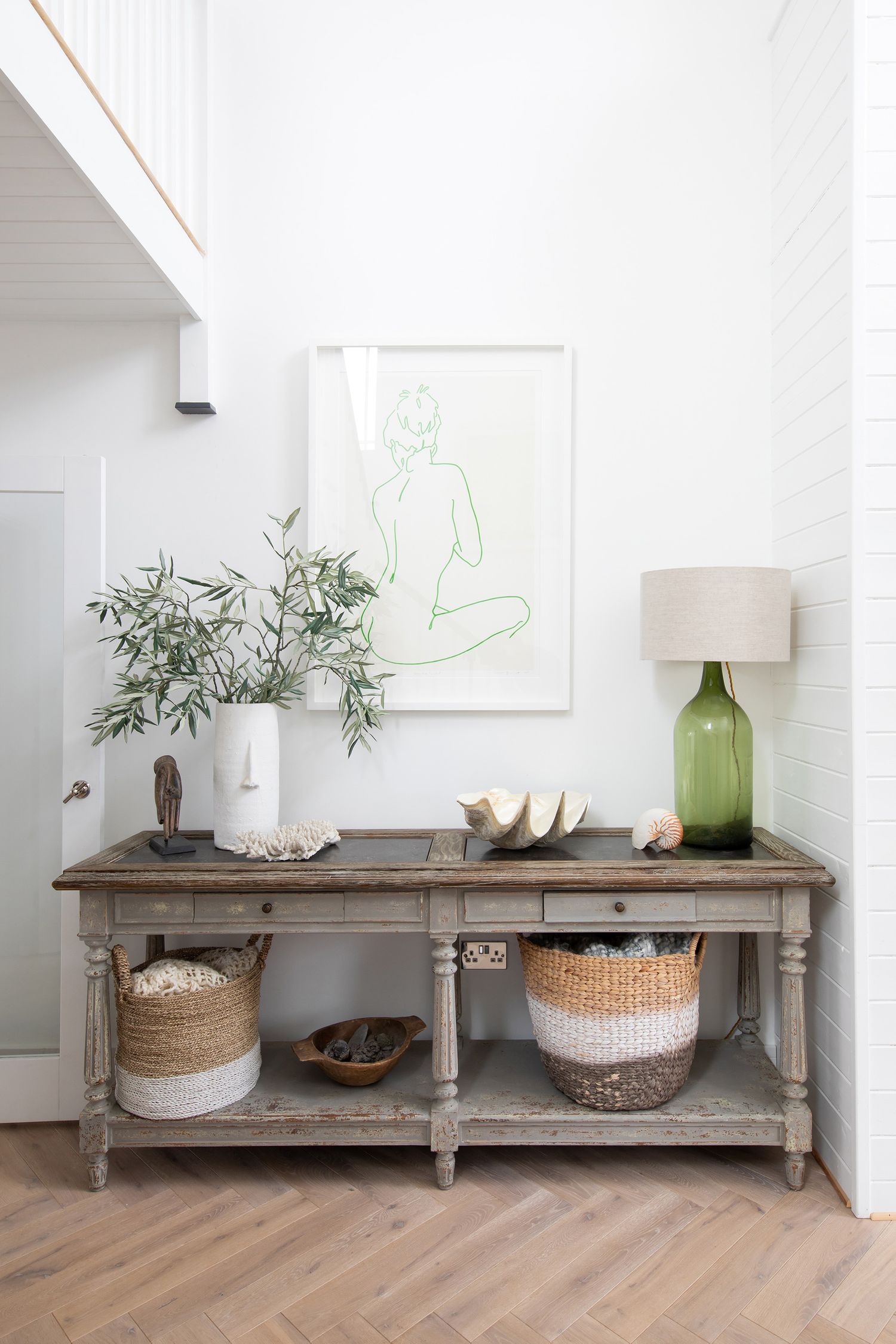 Rustic console table styled with green plants, baskets, and minimalist artwork in a bright white entryway.
