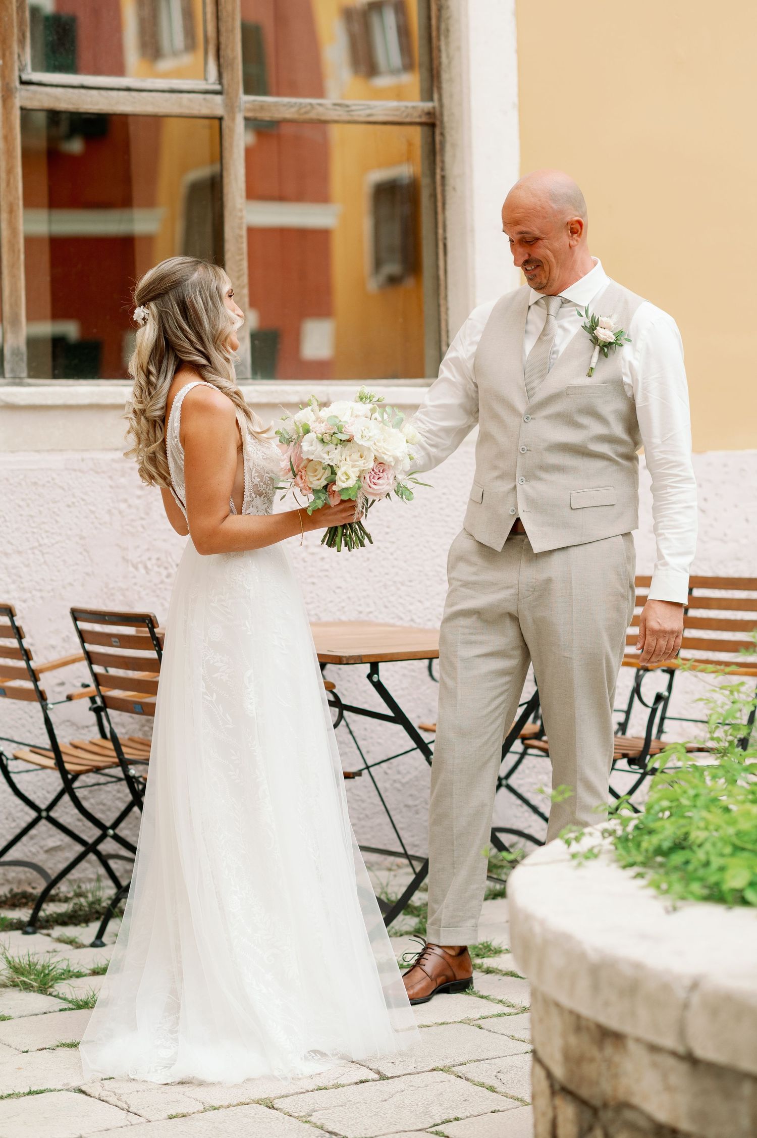 Couple shares first look moment in courtyard with bistro chairs and stone wall backdrop.
