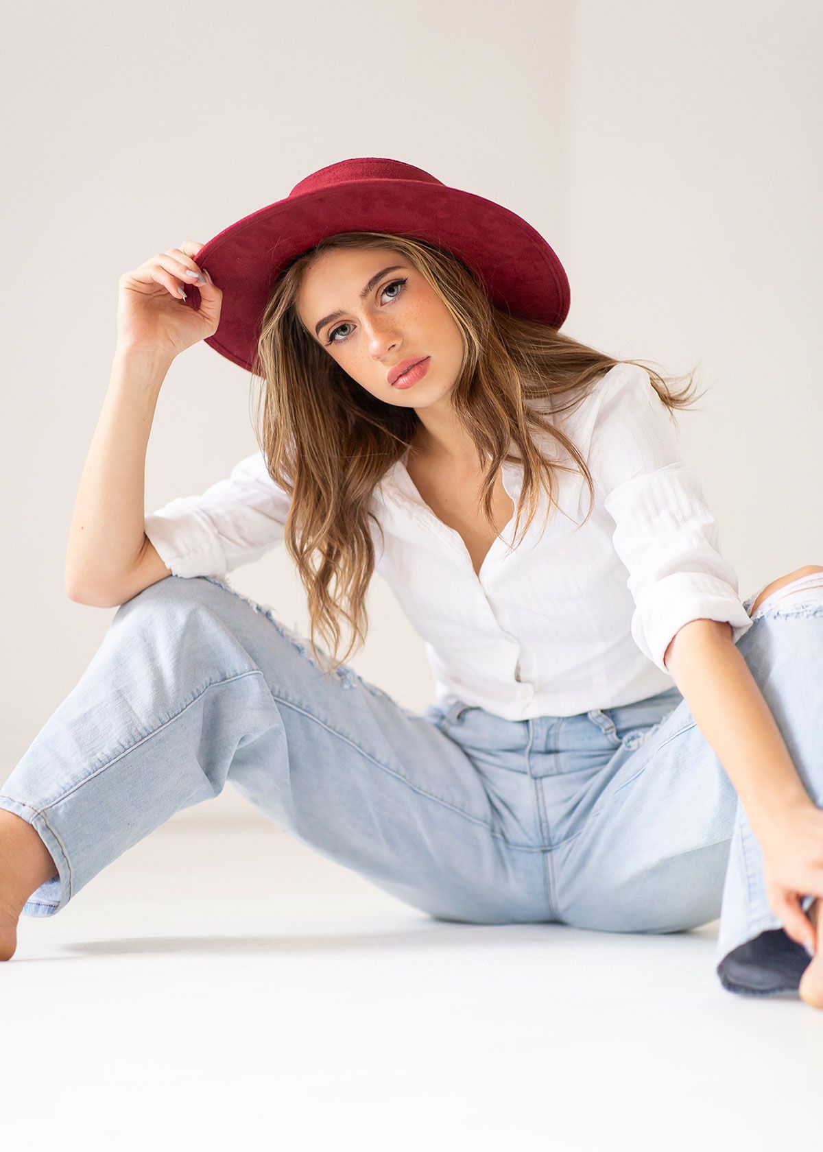 Woman in casual attire with burgundy hat, white shirt and light jeans sitting against white background.