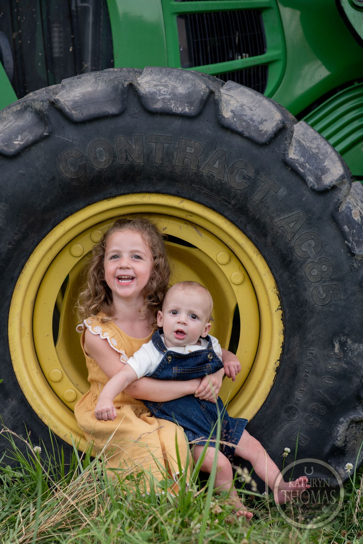 Two young children sit together inside a large yellow tractor wheel outdoors.