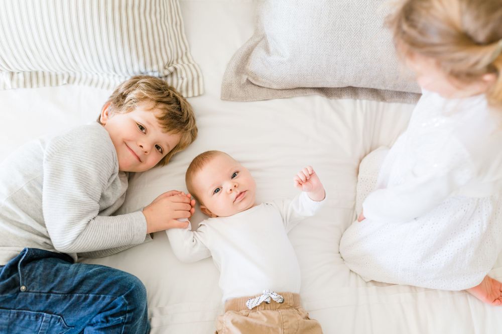 Two young siblings lying on a bed with their baby sibling, looking at each other with soft smiles in a cozy, light-filled setting.