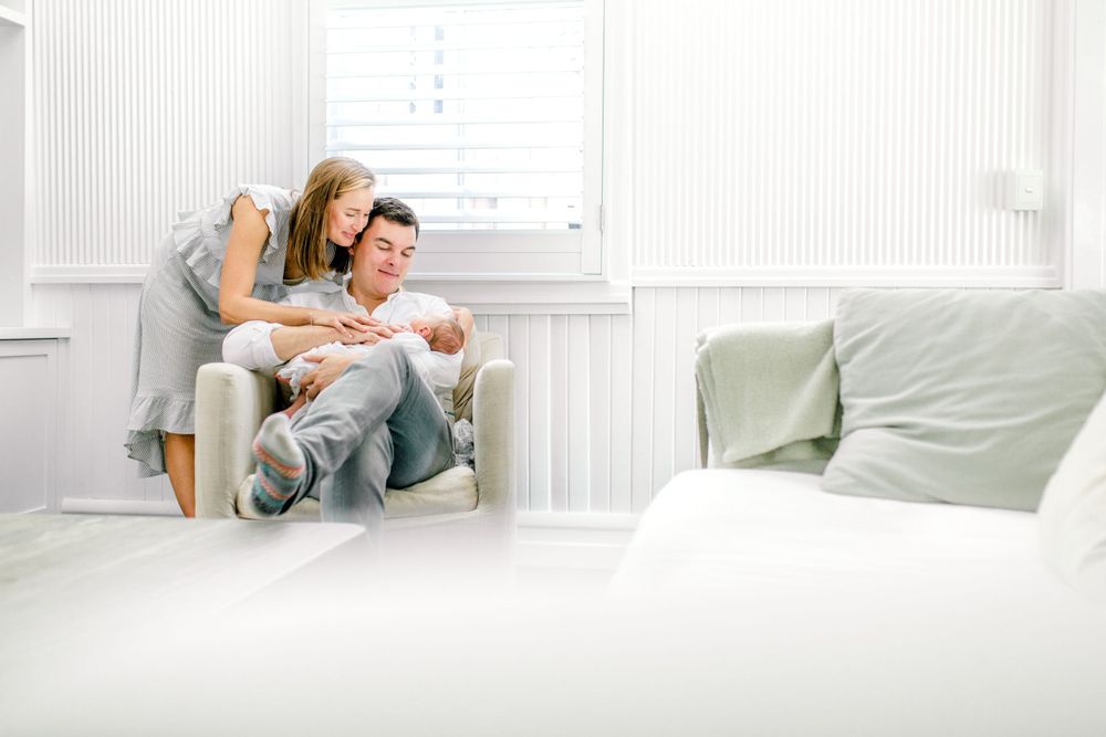 Mum and Dad sitting on a chair looking at their newborn baby in a light filled living room.