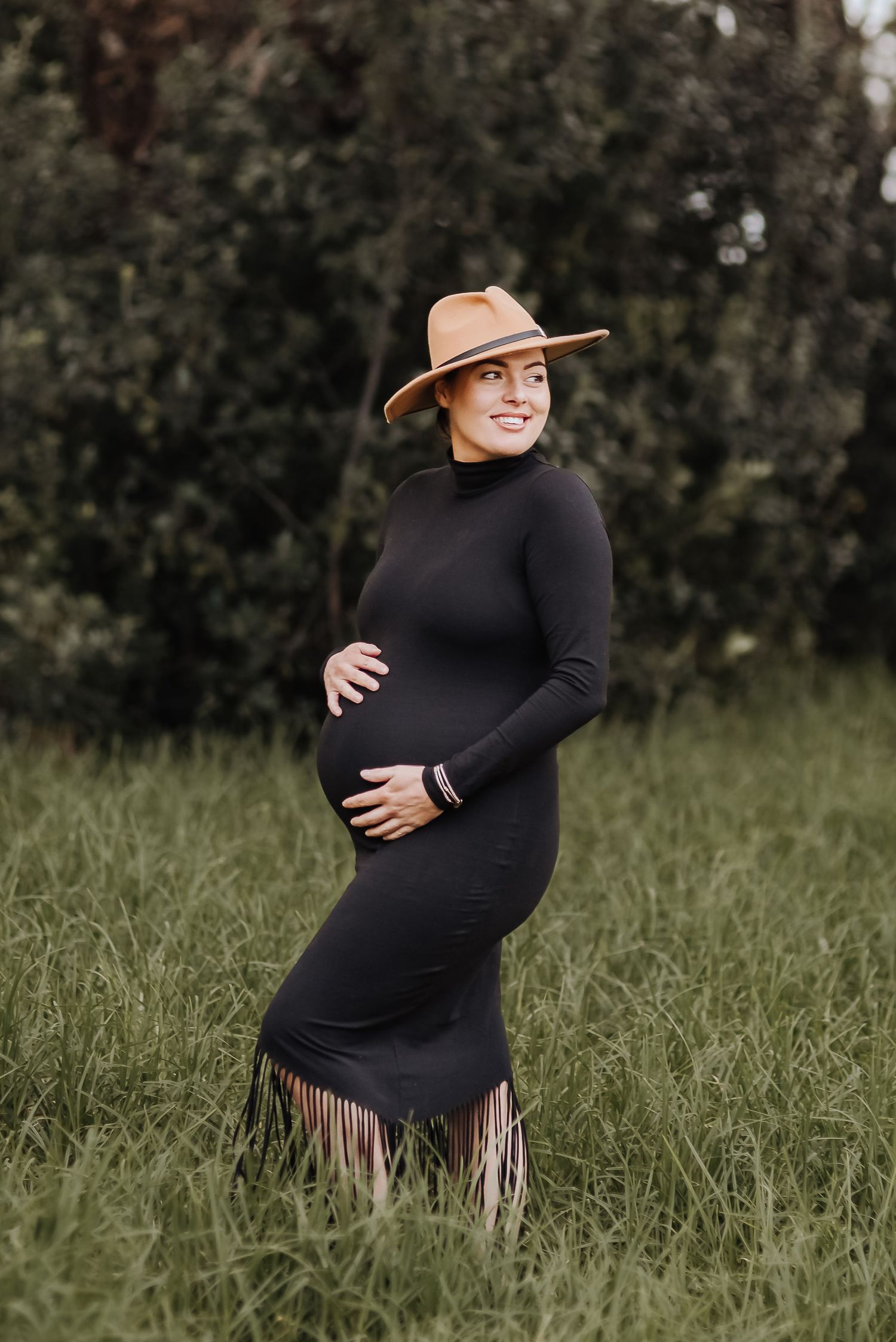 Maternity photoshoot in a field featuring a black dress with fringe and brown hat.