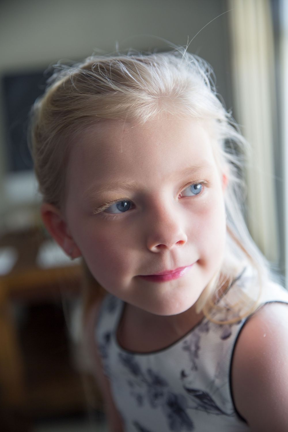 Young child in white dress poses near window with soft lighting.