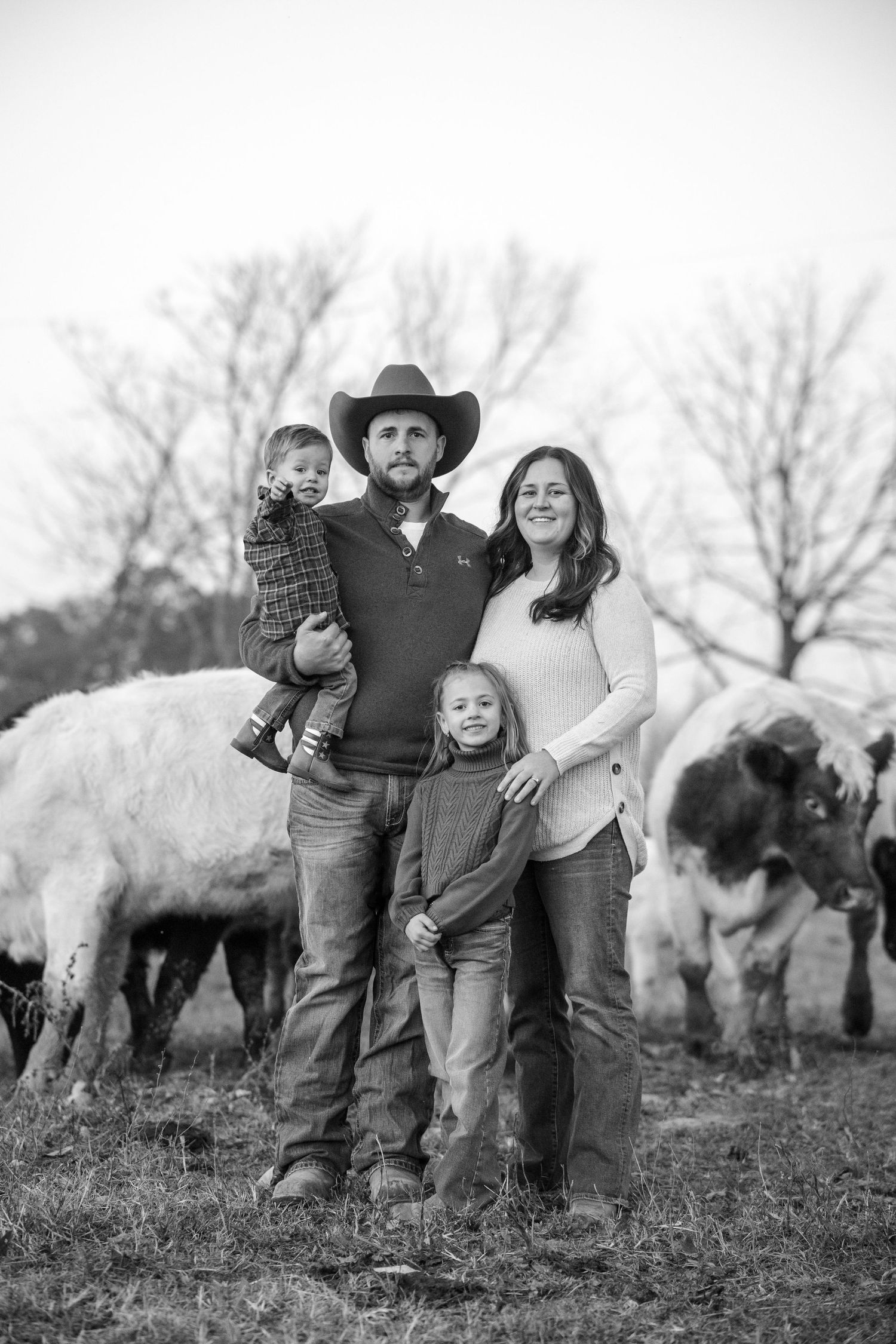 Black and white family portrait of couple with two young children standing among cattle in a field, man wearing cowboy hat.