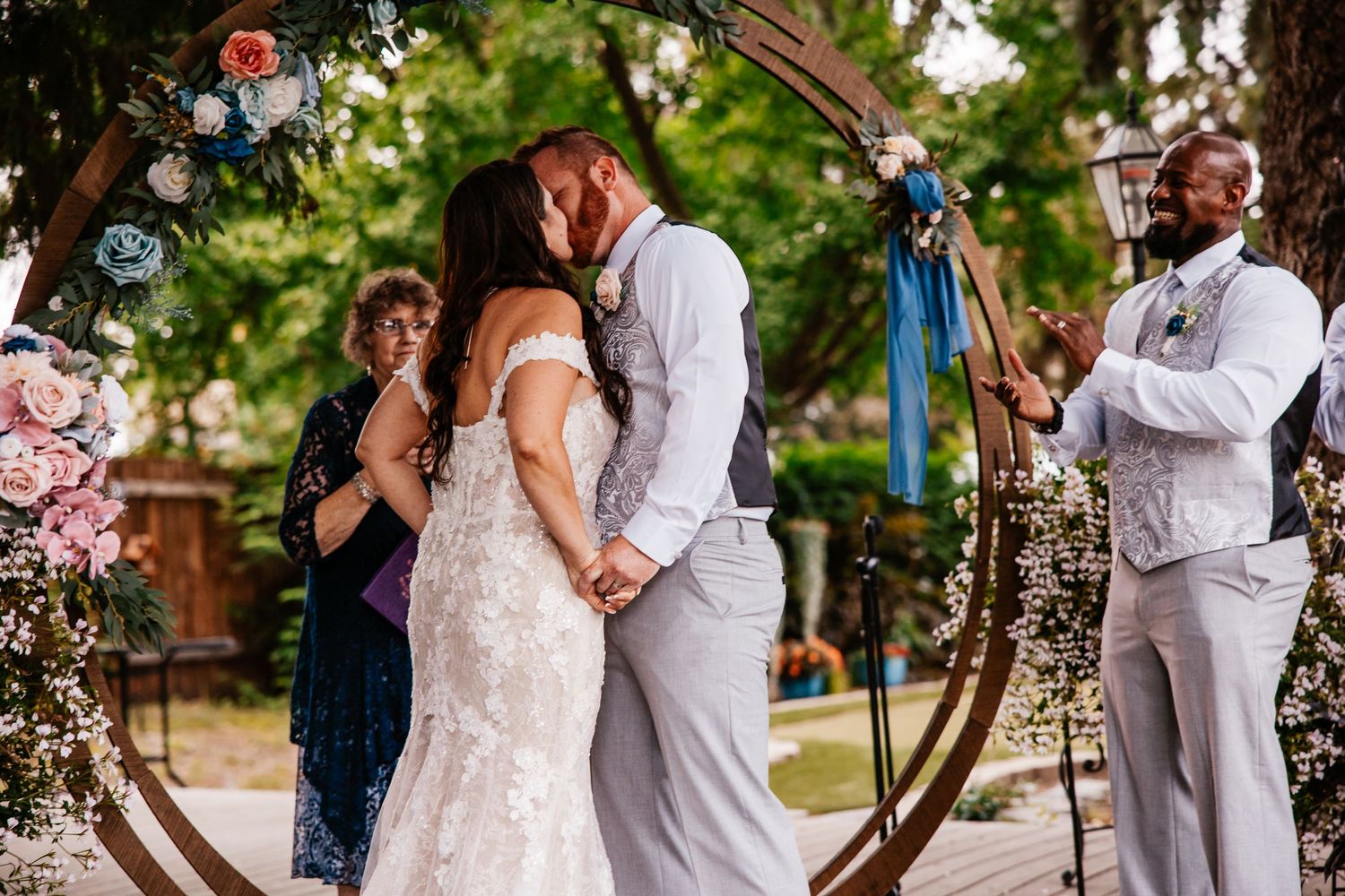 An outdoor wedding ceremony under an archway decorated with flowers and blue ribbons.