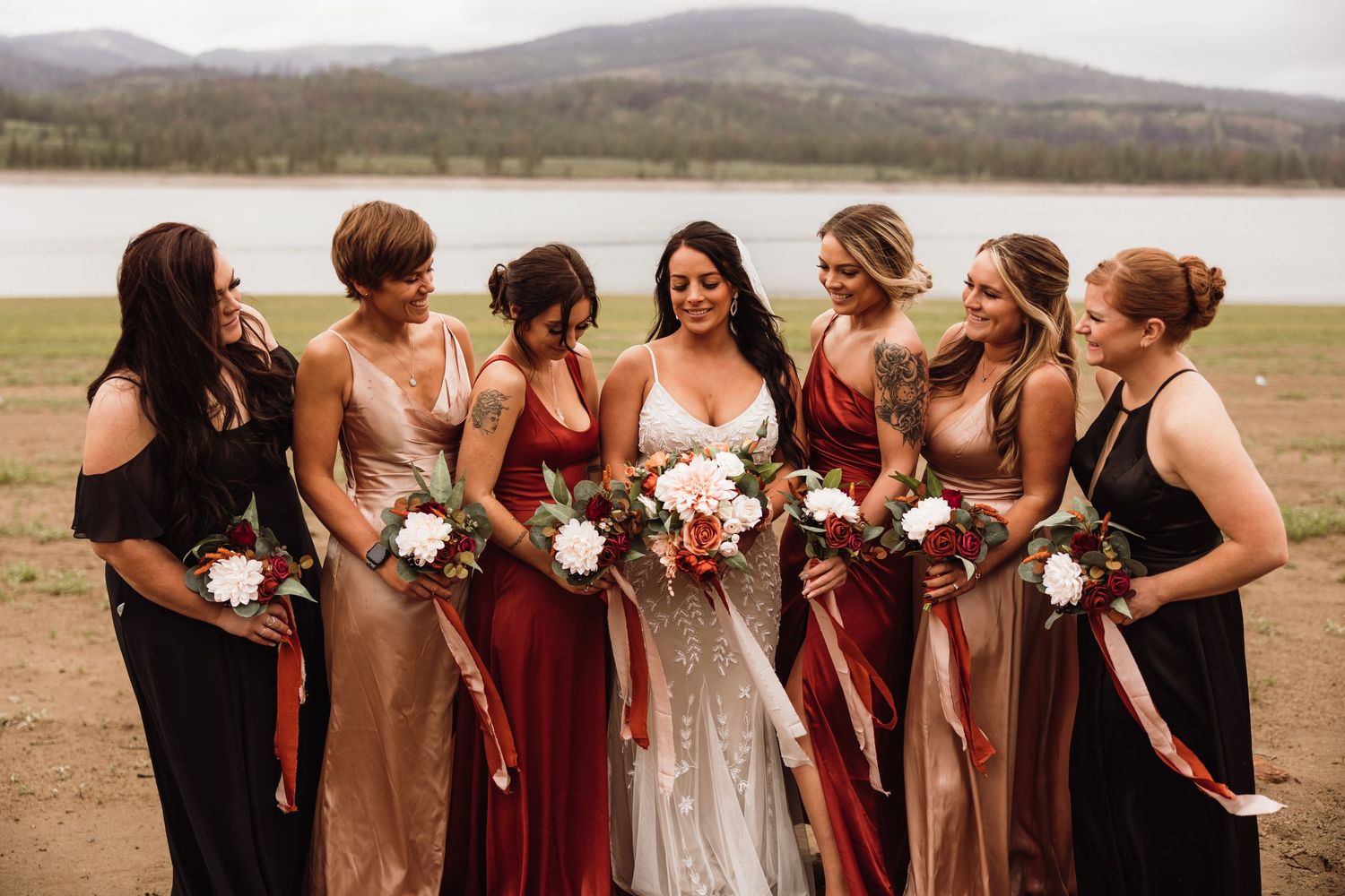 Wedding party in mix of burgundy and neutral dresses pose by lake with mountains in background.