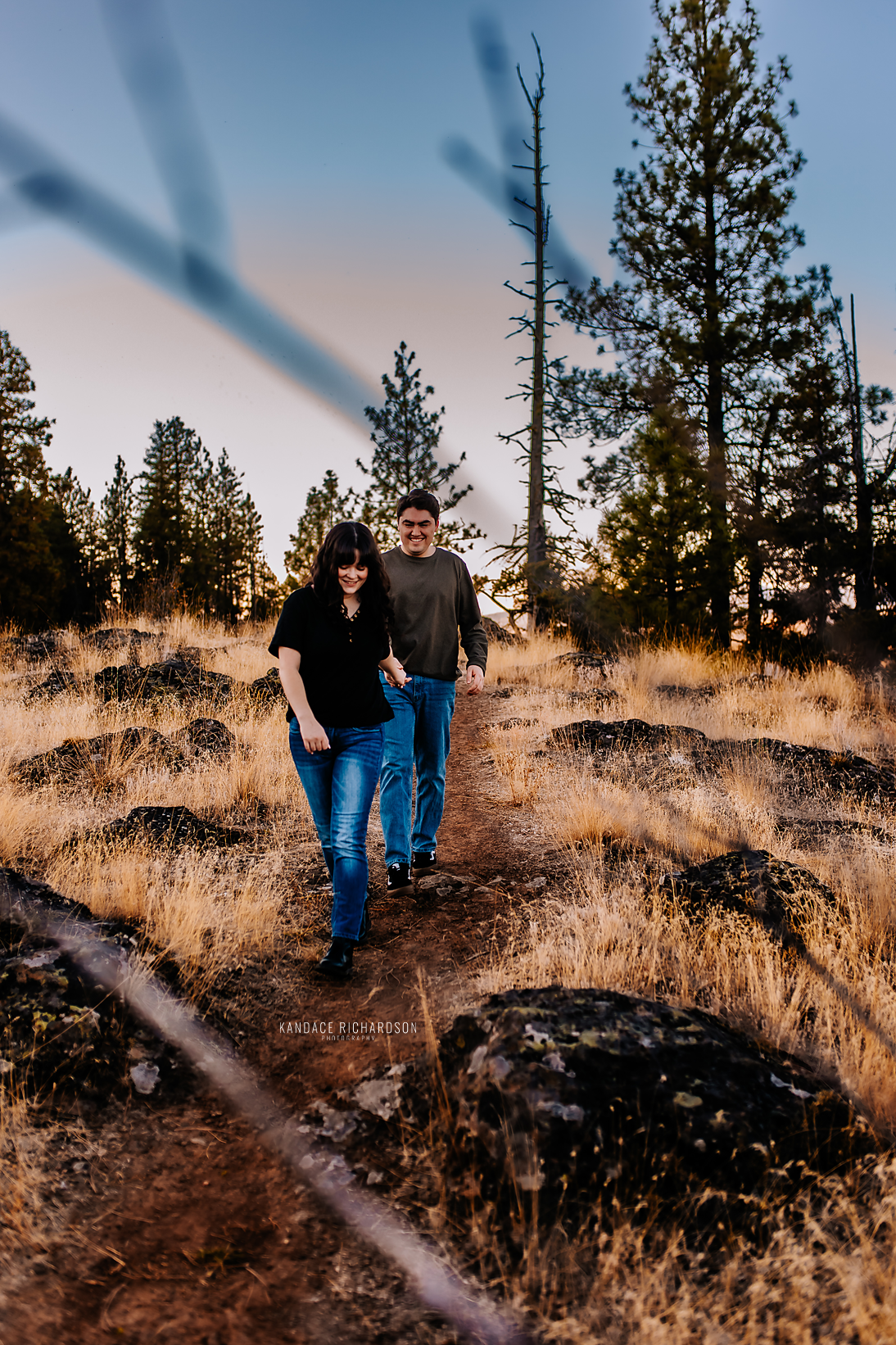 Couple walking through rocky terrain at dusk with pine trees silhouetted against pastel sky.