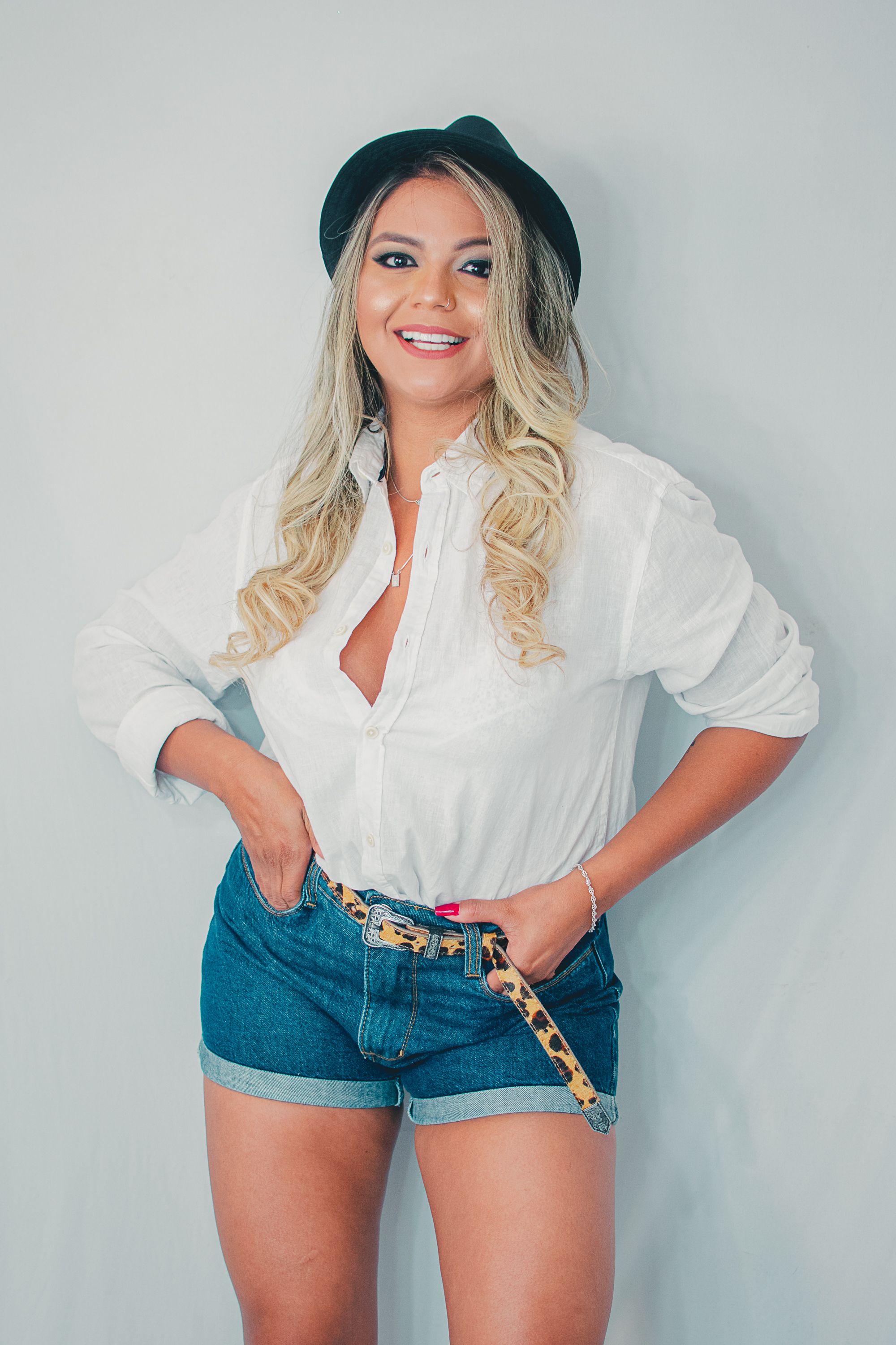 Model poses in denim shorts and white button-down shirt against light grey background.