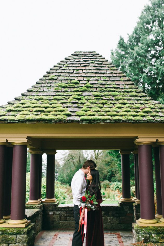 A romantic couple embraces under a moss-covered gazebo with stone pillars in a garden setting.