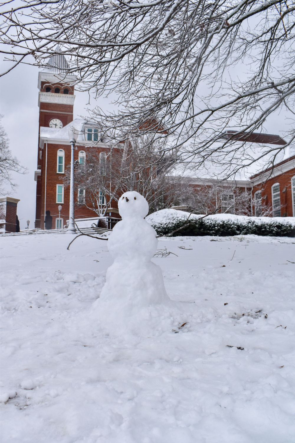 Snowman stands in front of a brick clock tower building on a snowy winter day.