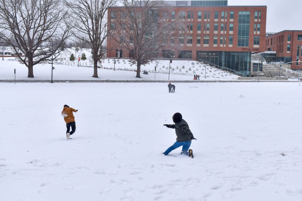 Two people playing in the snow on a winter day with red brick buildings in the background.