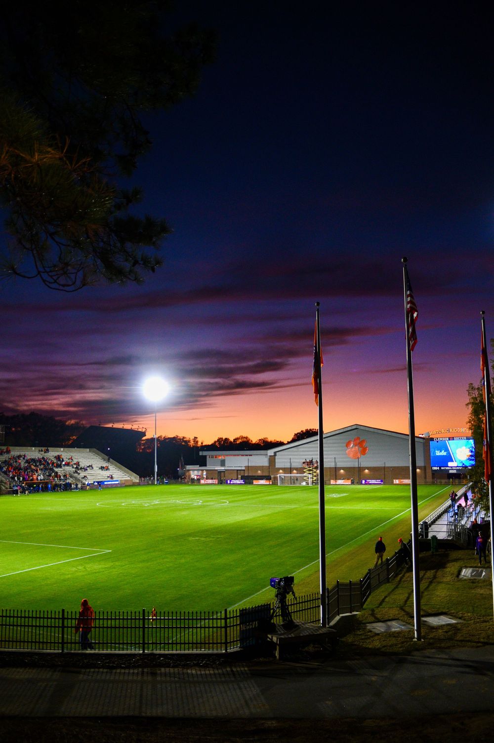 Illuminated sports field at dusk with flag poles and purple sunset sky.