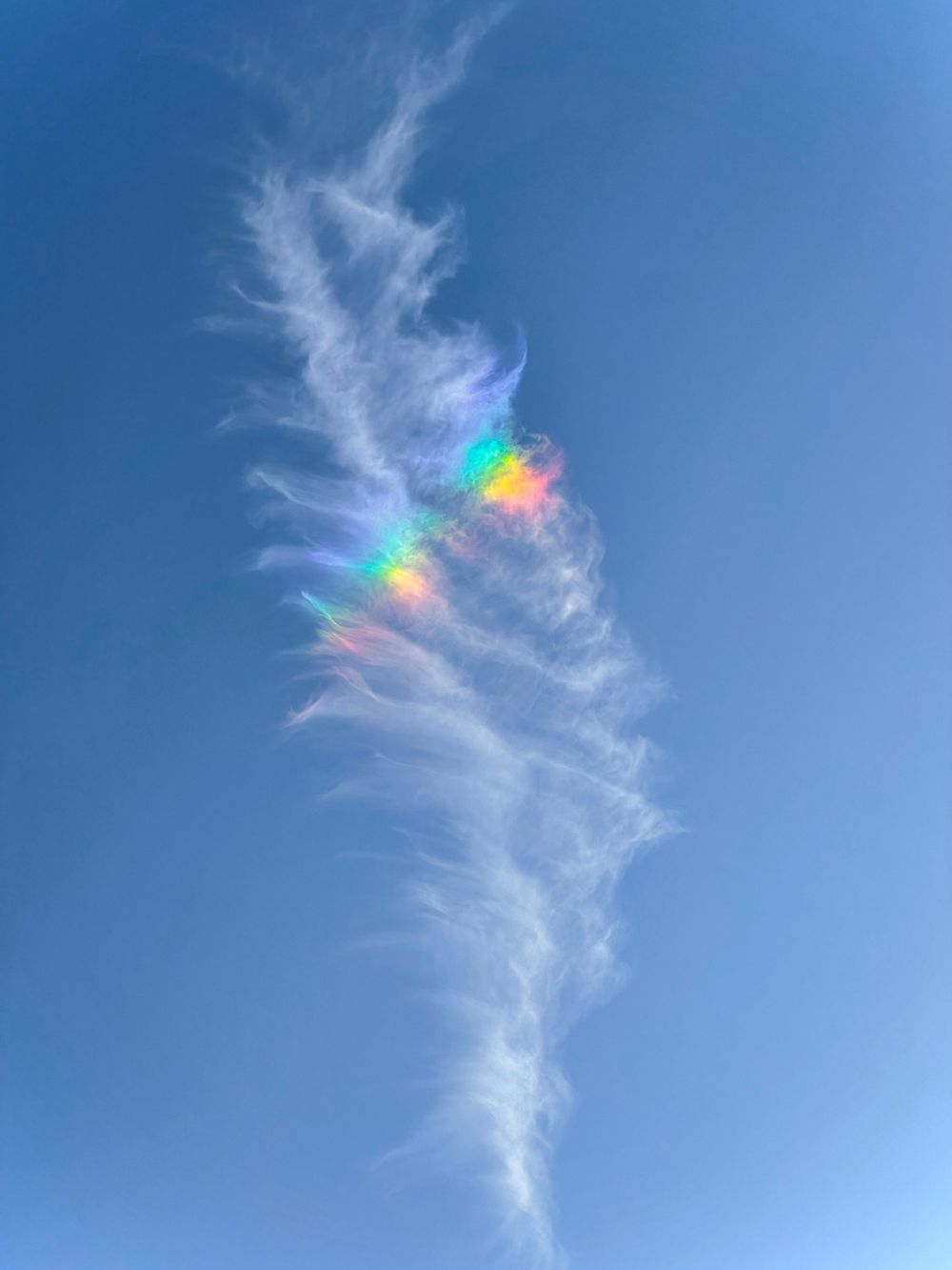 Wispy cloud formation with rainbow iridescence against a bright blue sky.