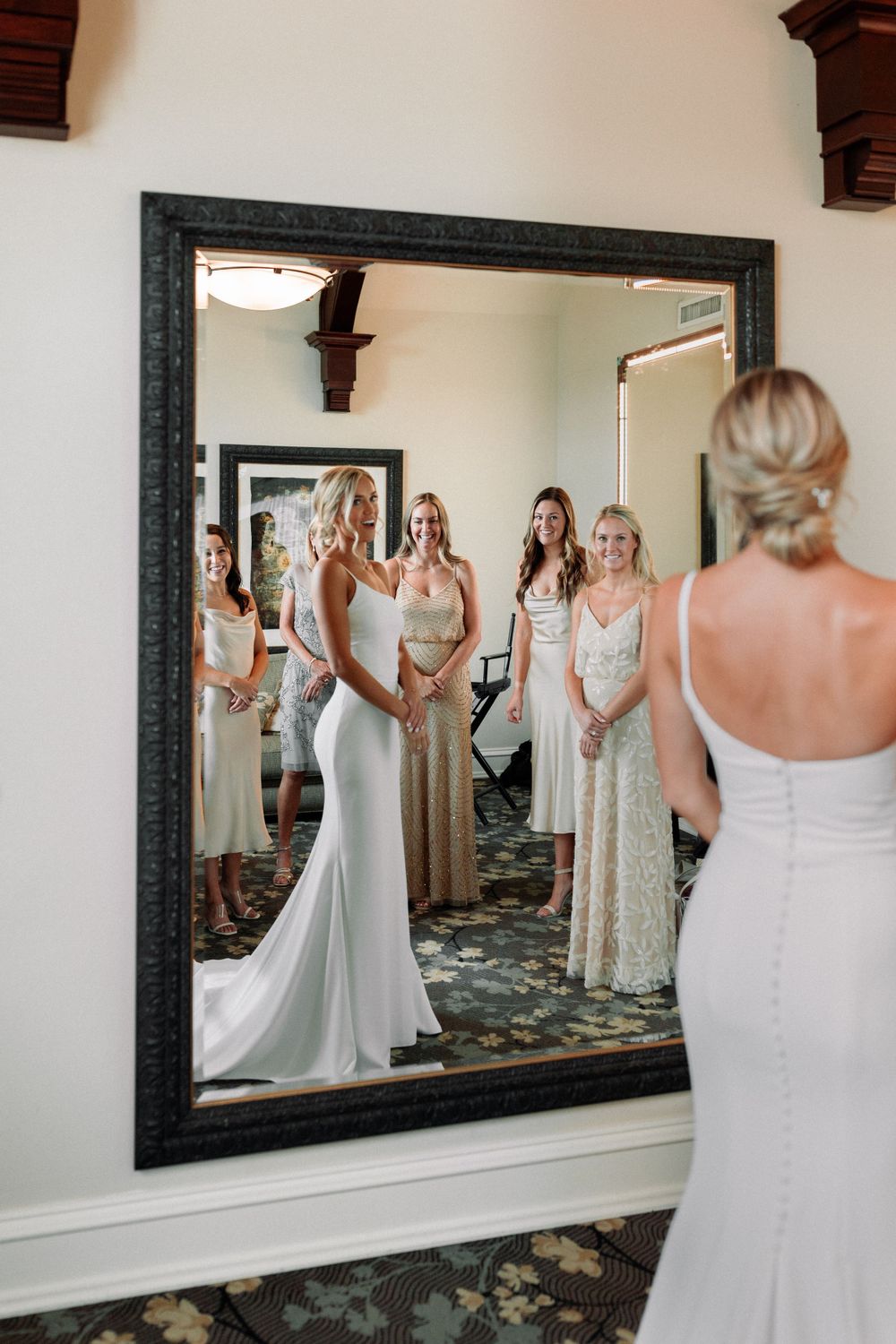 Bridal party gathered around large mirror watching bride in white gown before ceremony.