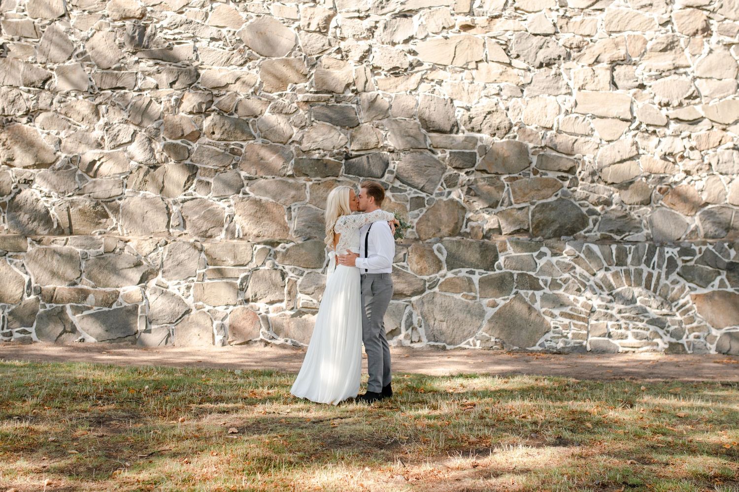 Wedding couple stands together against rustic stone wall backdrop in sunlight.
