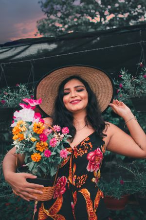 Garden scene with someone in a floral dress and straw hat holding a colorful flower arrangement.