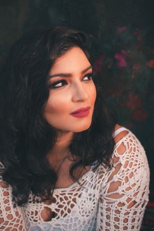 Close-up portrait featuring someone in a white lace top against a dark floral background.