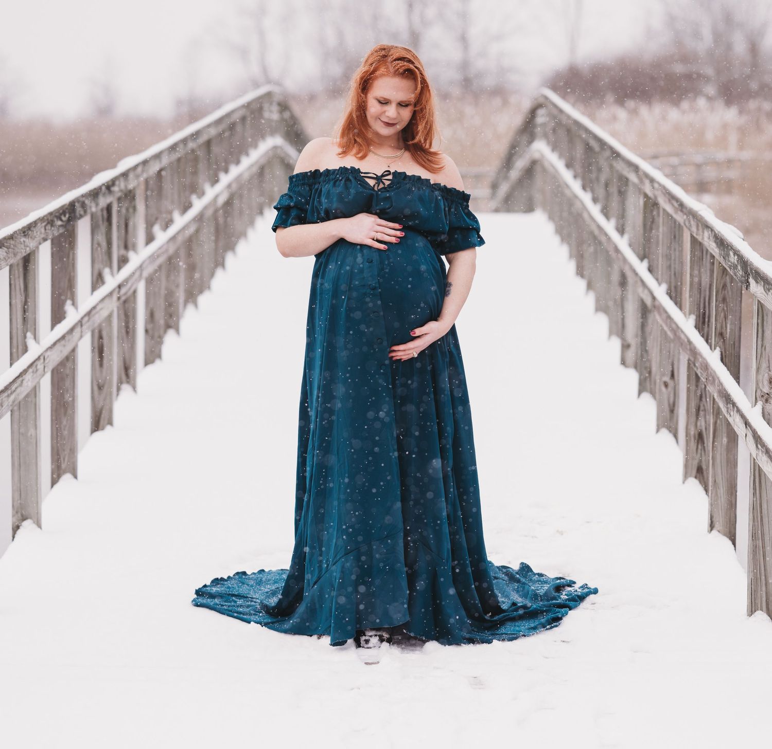 Maternity portrait on a snowy bridge featuring a flowing teal velvet gown.