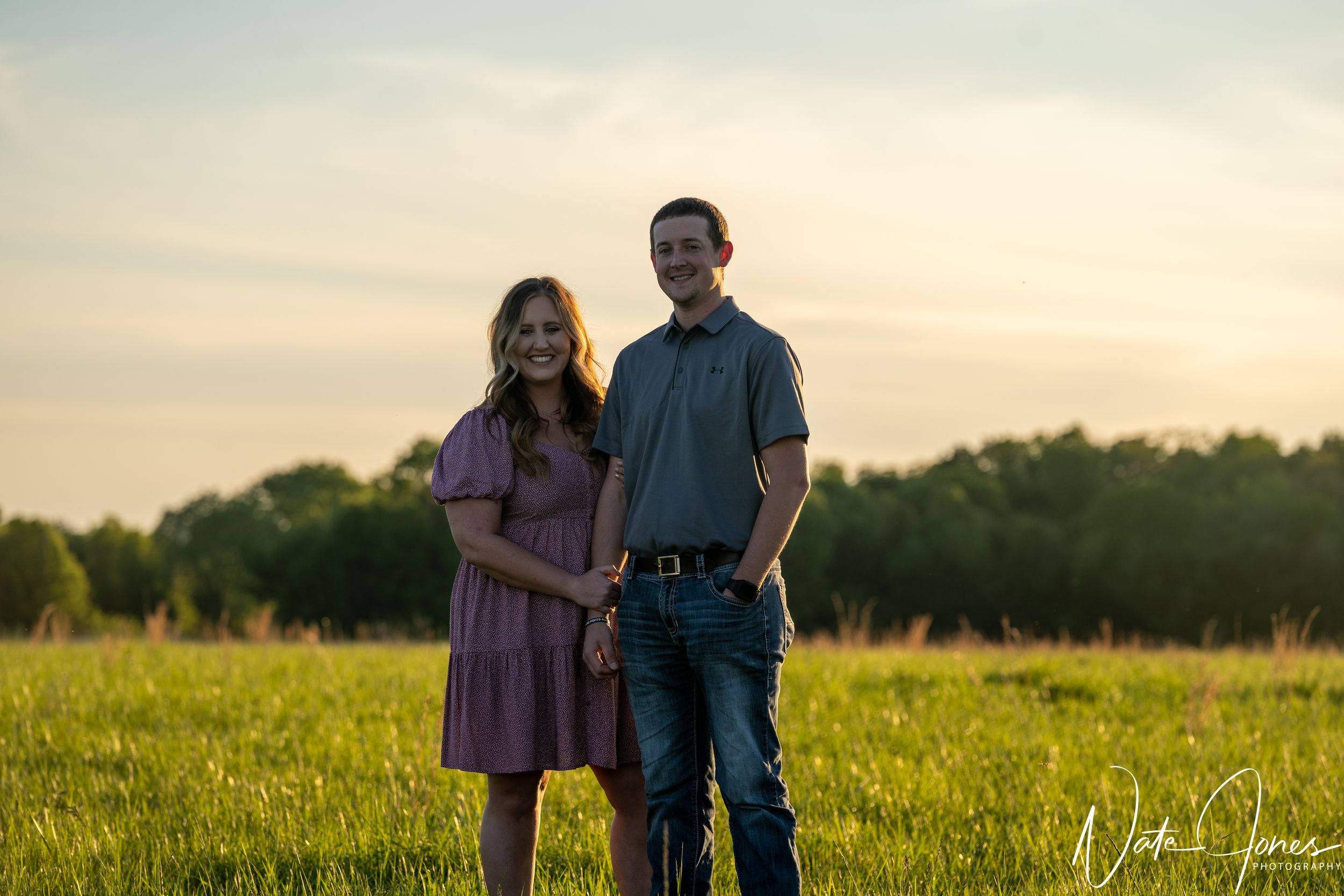 A couple poses in a grassy field at sunset with warm golden lighting.