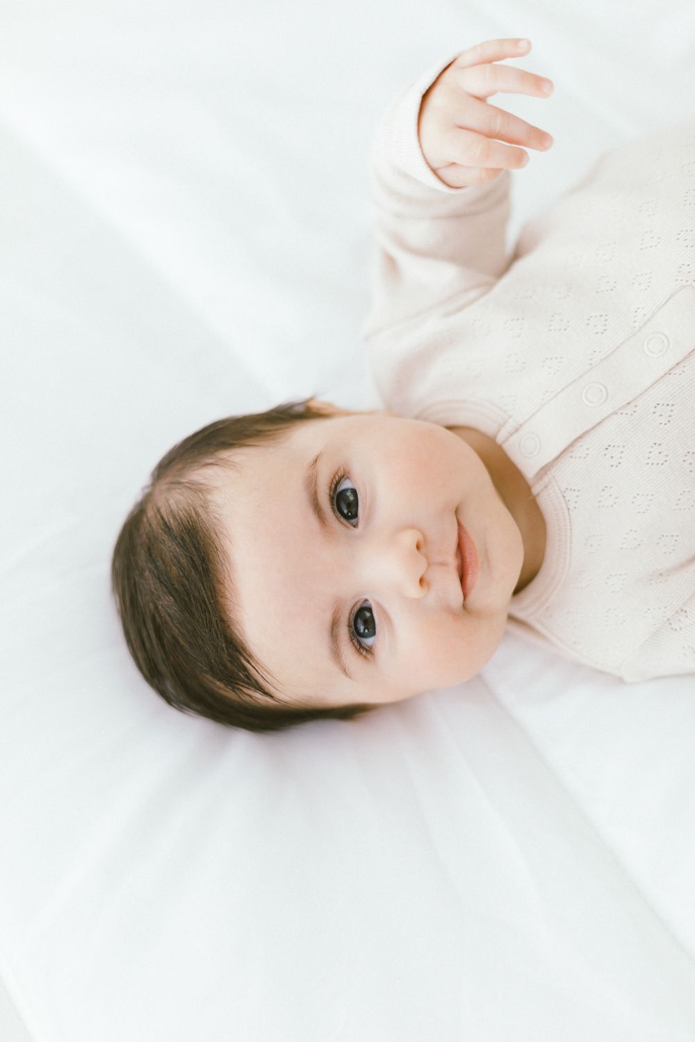 Baby in ivory sweater lying on white sheets with bright eyes.