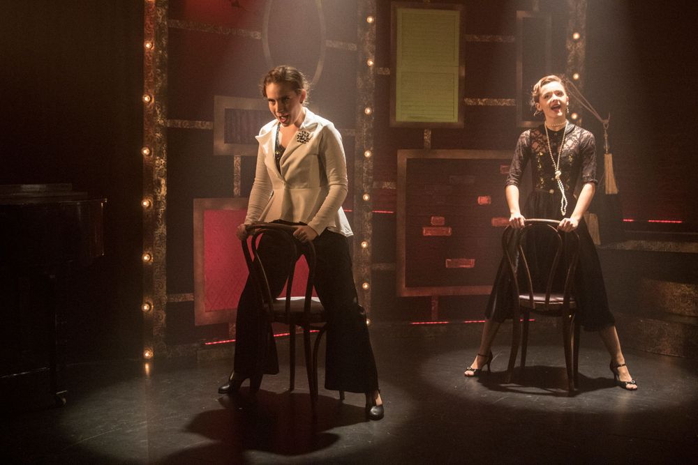 Two performers sit on bar stools under stage lighting during a theatrical performance.