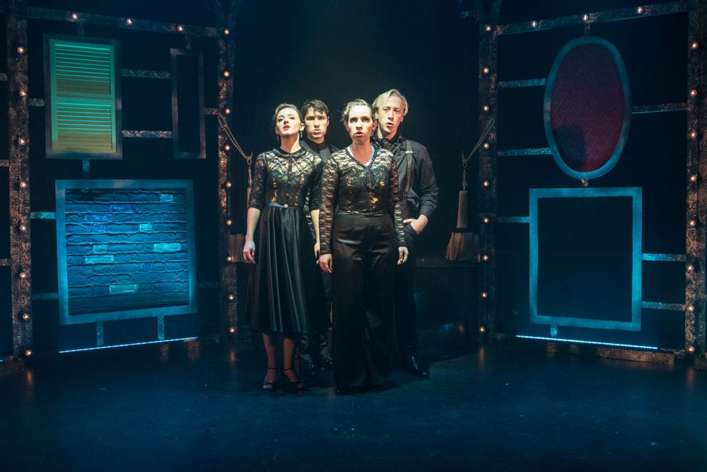 Performers stand posed in dramatic lighting on a theatrical set with glowing screens and string lights.