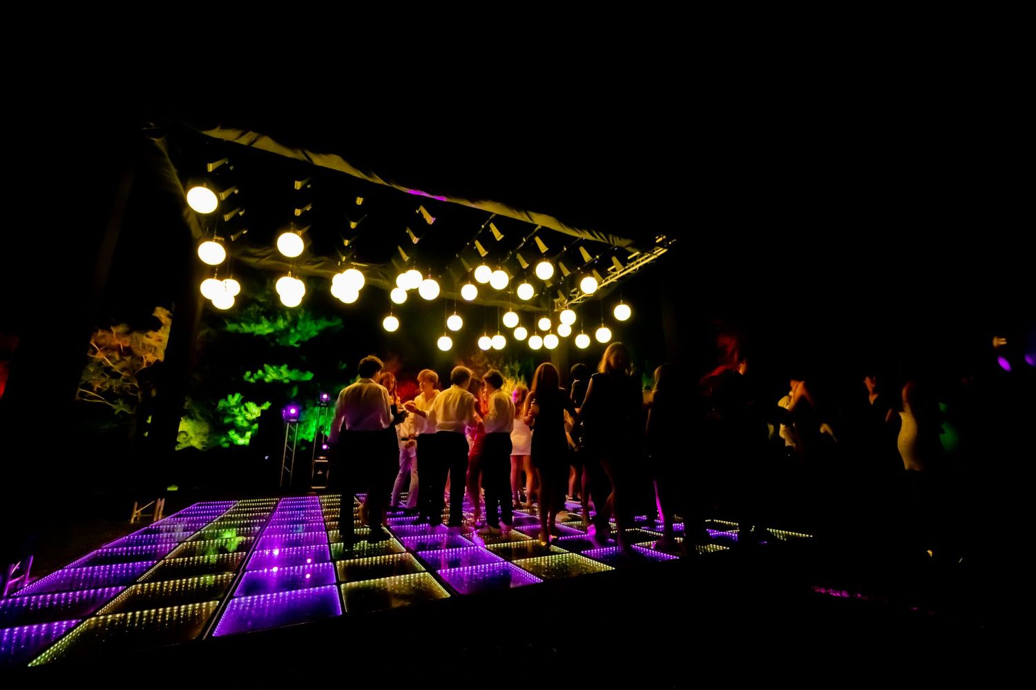 People dancing on colorful LED dance floor under string lights at outdoor night event.
