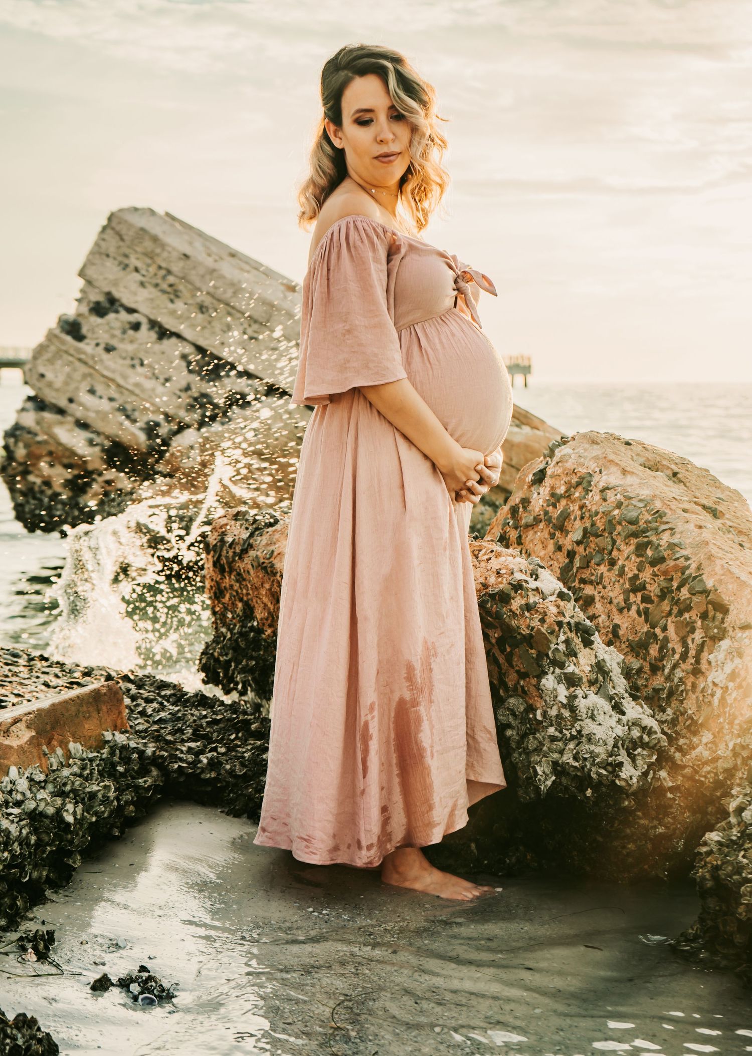 Outdoor Maternity Session at Ft. Desoto in St. Pete, FL; woman embracing her pregnancy belly near rocks in ocean