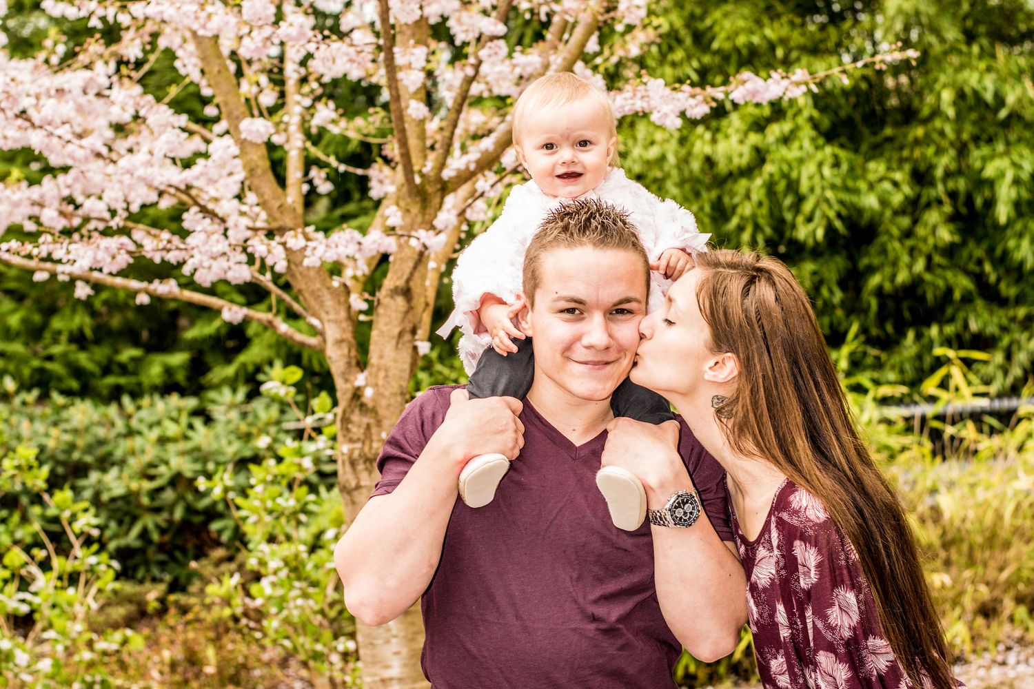 Happy couple enjoying a spring day outdoors with cherry blossoms and a baby on shoulders.