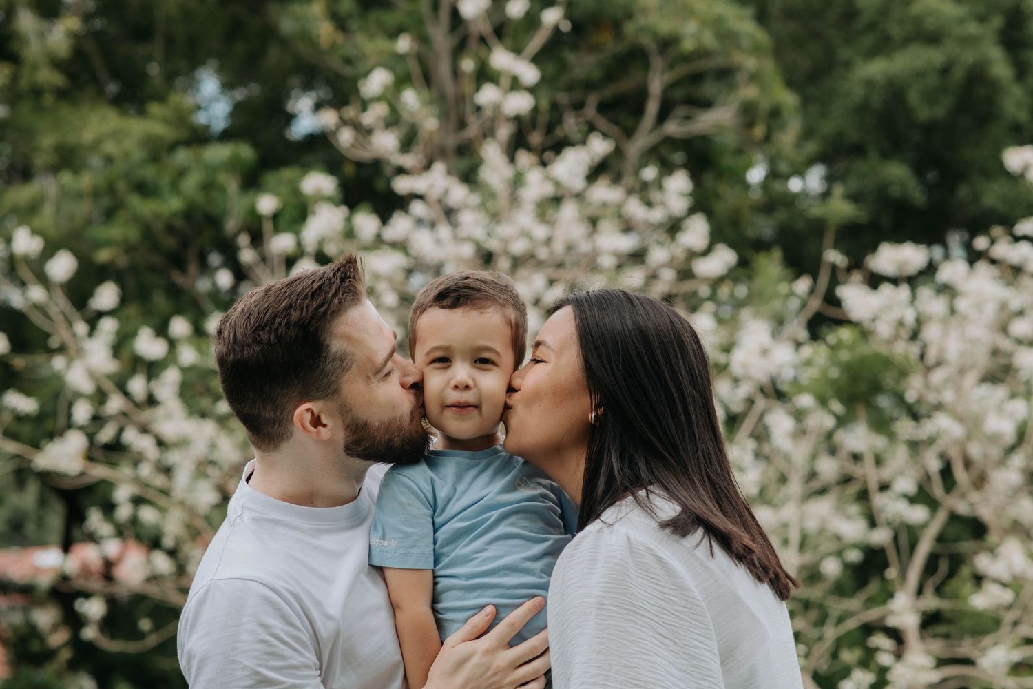 Loving parents kissing their toddler son against blooming spring trees.