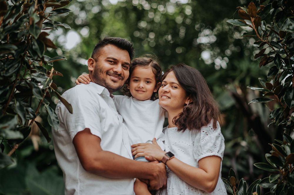 A family of three wearing white shares a joyful embrace among green foliage.