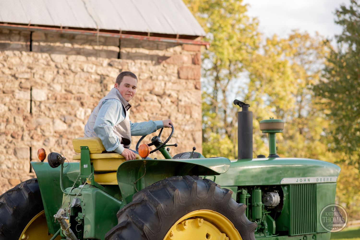 Person operates vintage John Deere tractor near stone building on farm.