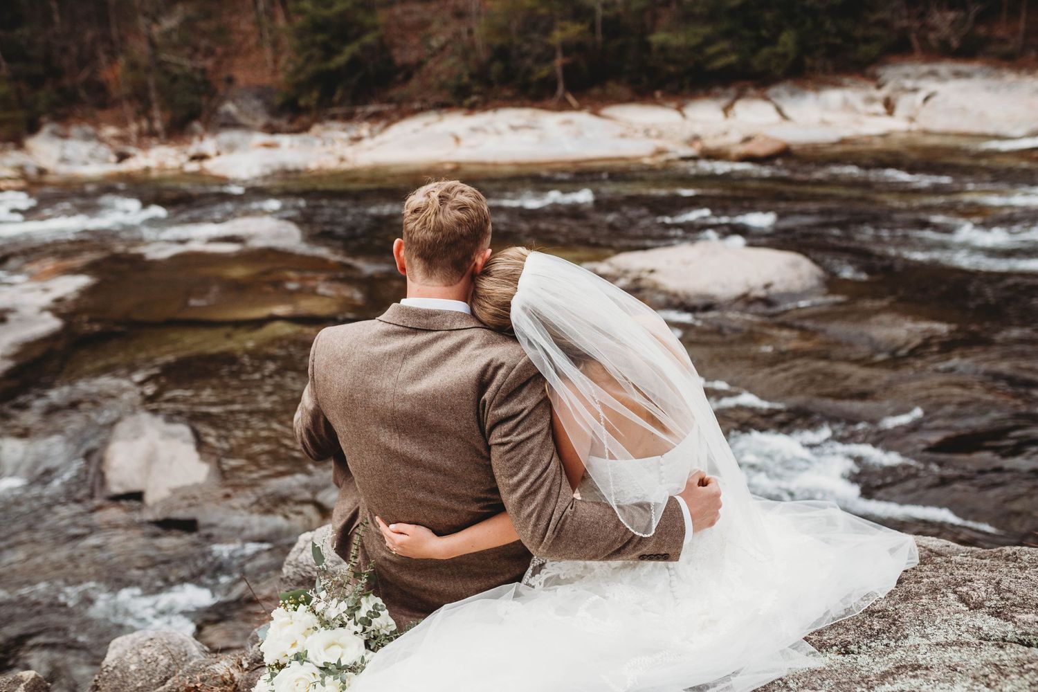 Couple in wedding attire sitting together on rocks overlooking a winter stream.