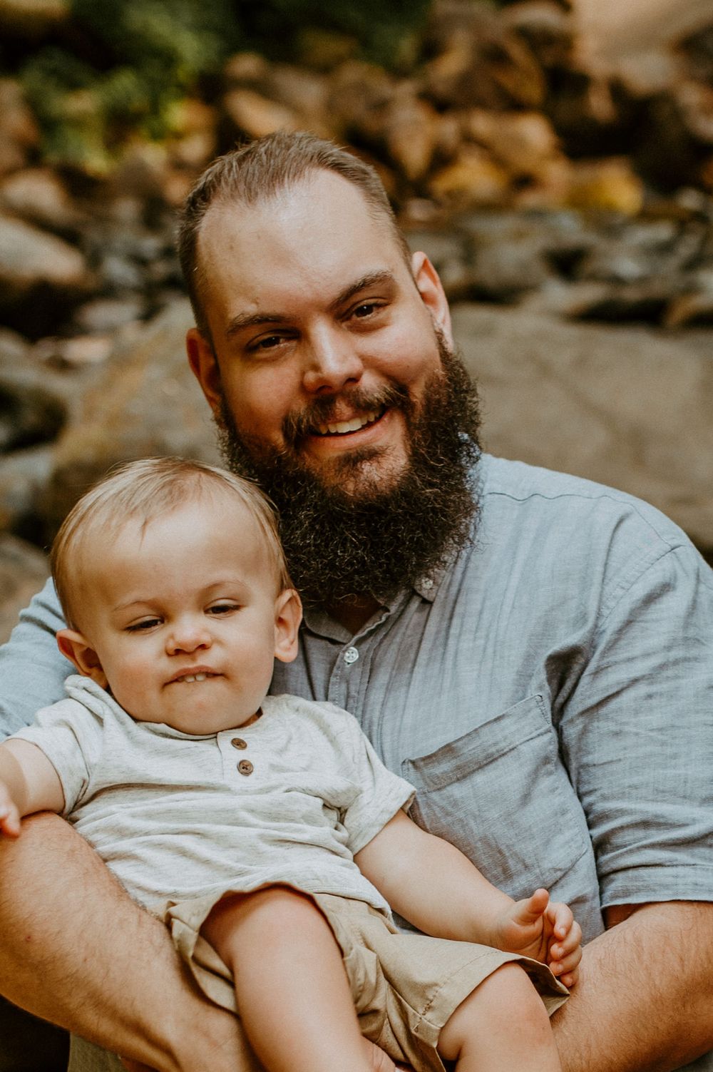 A bearded person holds a small child while sitting on rocks near a forest stream.