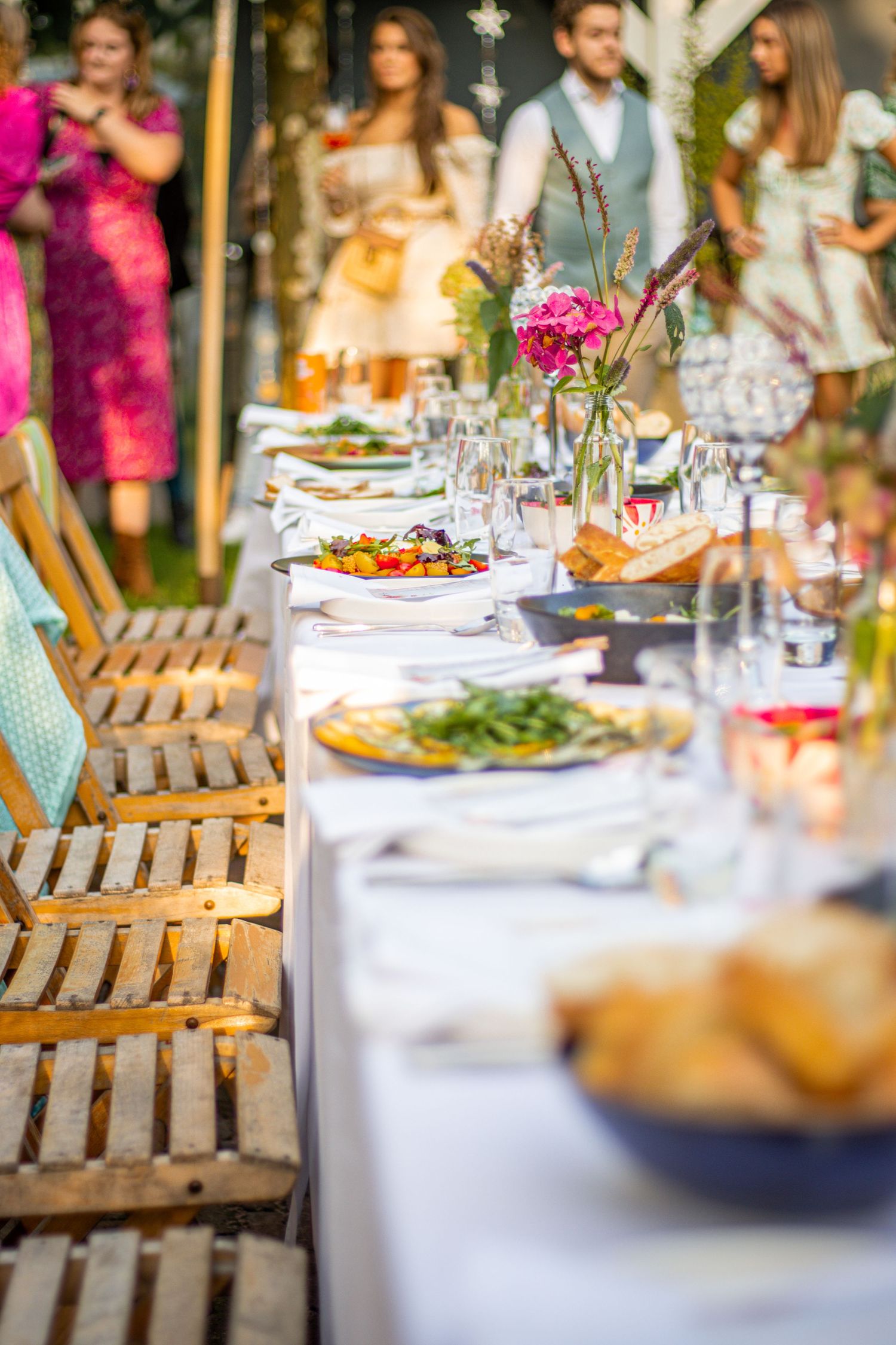 Zomerse tuintafel met Italiaans eten, houten stoelen en chique gasten in de achtergrond voor een feestelijke sfeer.